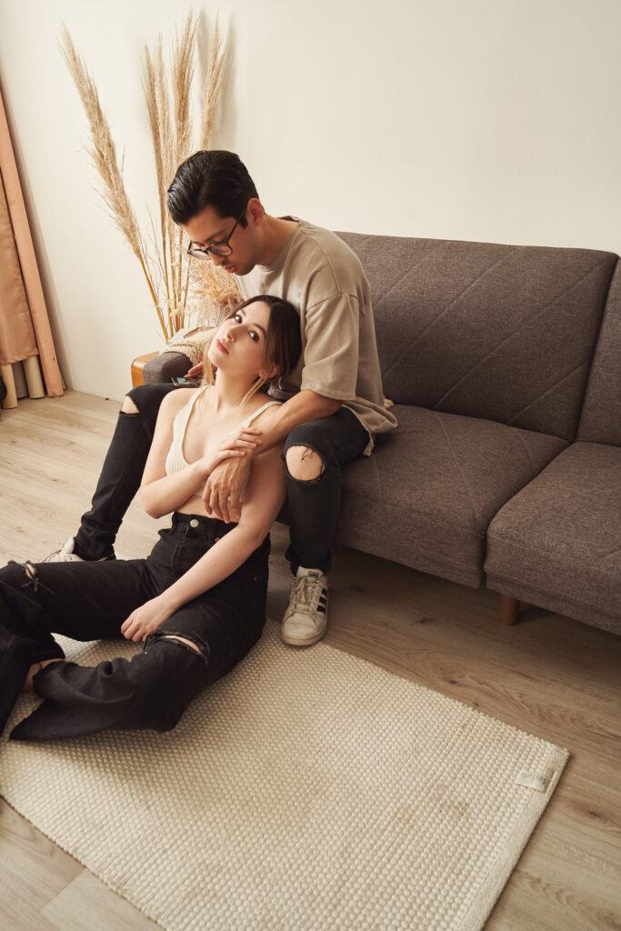 Gaby and Me a couple share a tender moment in a minimalist living room. Gaby sits on the floor leaning against I sits atop a sofa.
